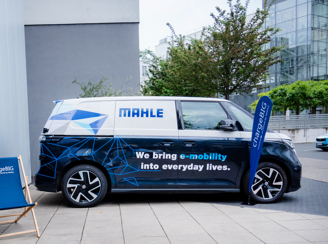 Ein Elektrofahrzeug von MAHLE chargeBIGmit der Aufschrift „We bring e-mobility into everyday lives“ ist vor einem modernen Gebäude geparkt.