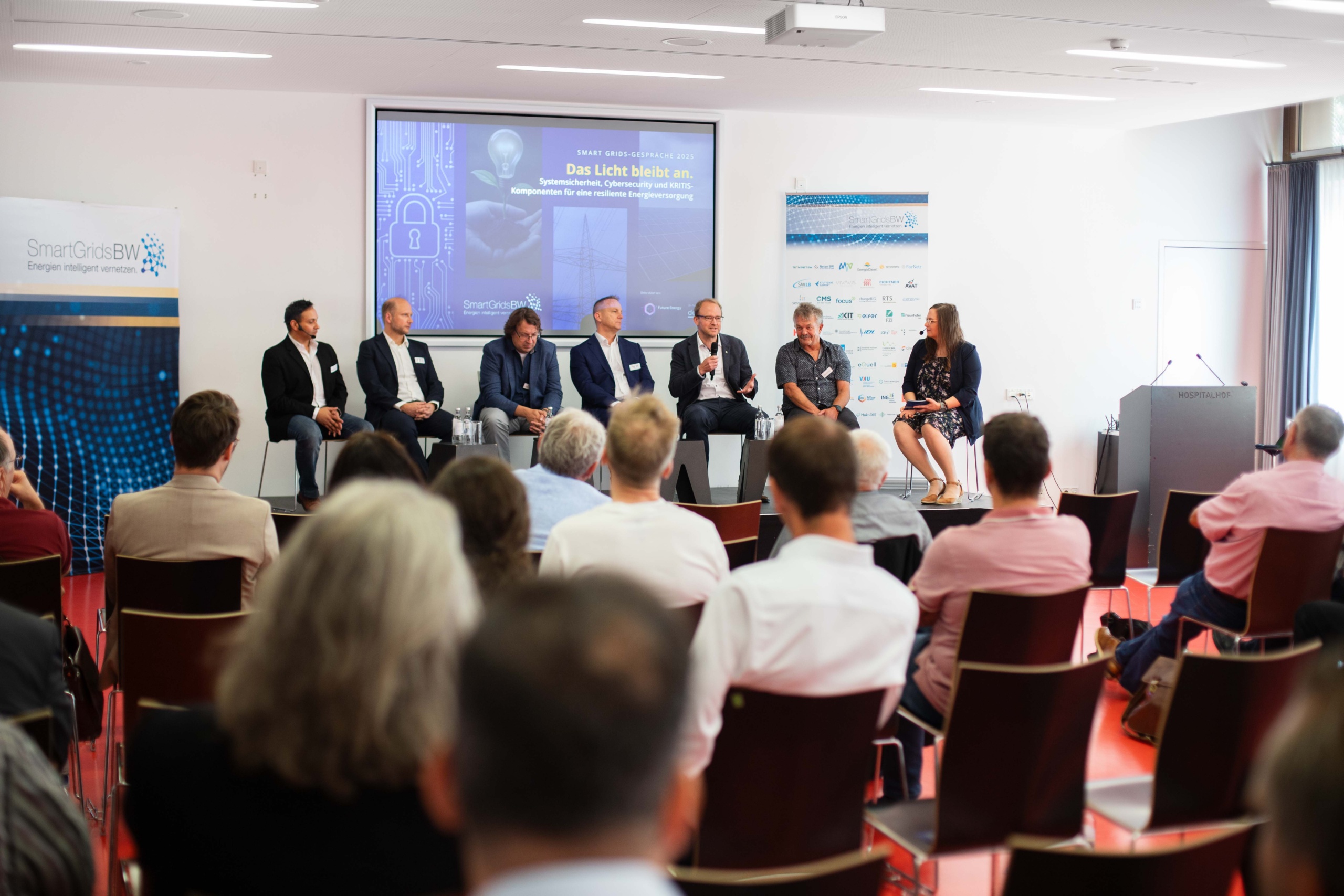 Foto: SmartGridsBW (Lea-Philine Beyer) Smart Grids-Gespräche zum Thema Resilienz: Wie wir kritische Infrastrukturen sichern können. Mehrere Menschen sitzen auf der Bühne und unterhalten sich. Einer spricht in ein Mikrophon.