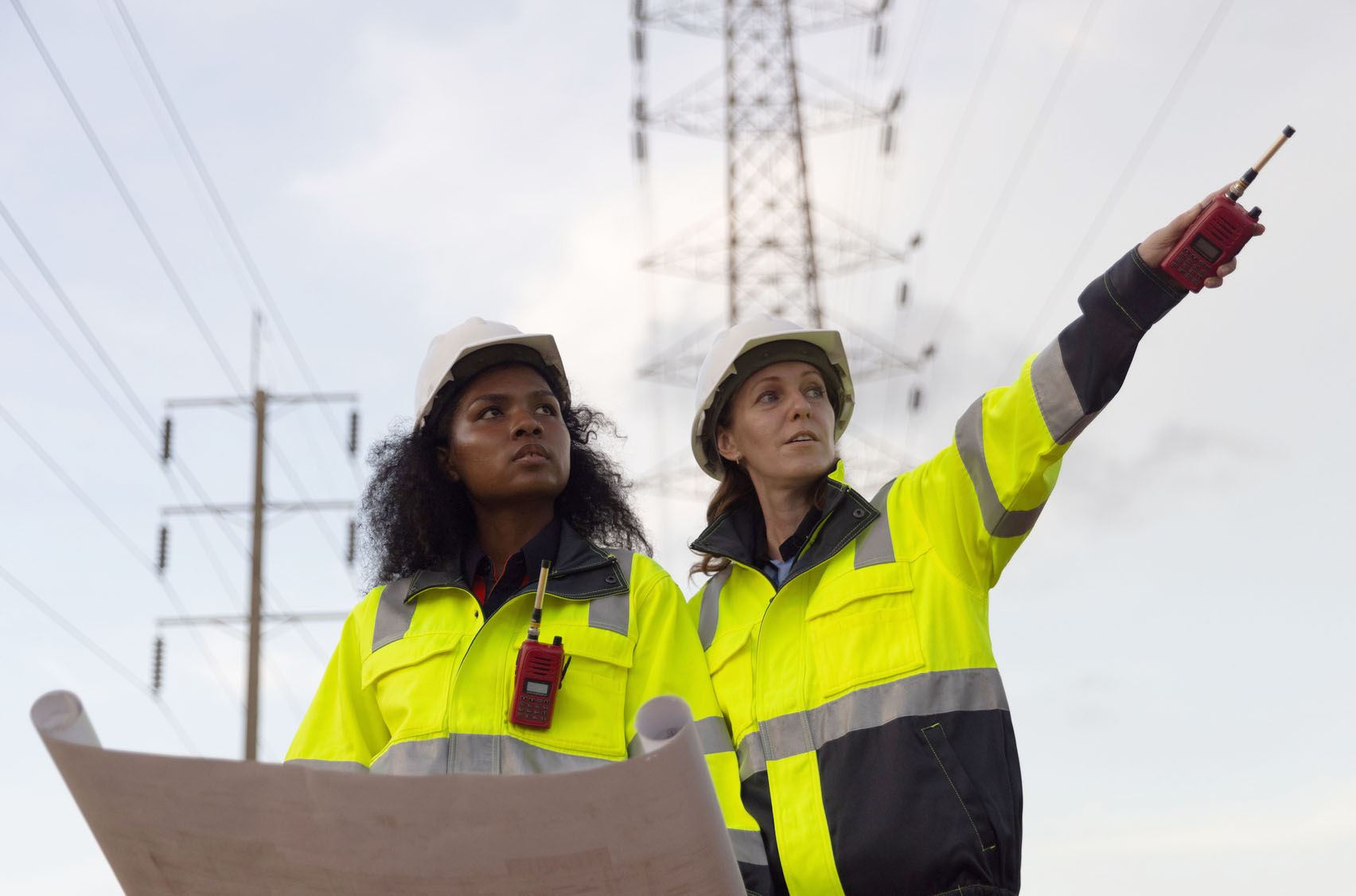Diverse two women electrical engineers holding blueprints looking confident with power line towers to project planning work producing electrical energy high voltage, energy concept.